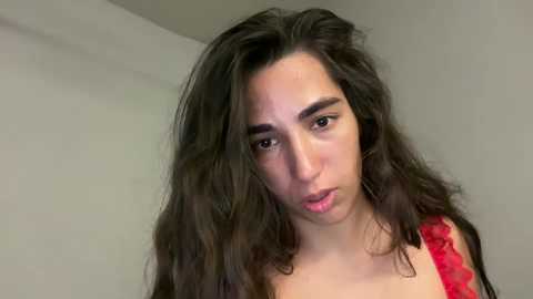 Media: Video of a young woman with long, wavy dark brown hair and fair skin, wearing a red lace top, looking introspective against a plain, off-white background.