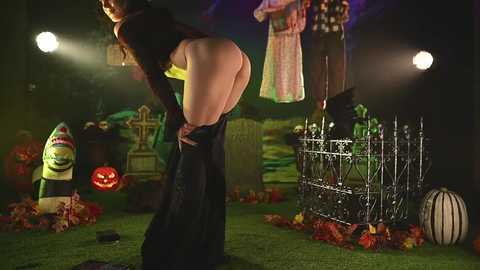 Media: Video of a woman in black lingerie, bending over, in a Halloween-themed graveyard. Background includes jack-o'-lanterns, a witch, and a coffin.