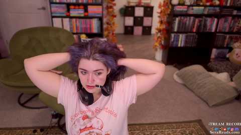 Media: Video of a woman with dark hair, wearing headphones, standing in a room with a green chair, bookshelves, and a cozy carpet.