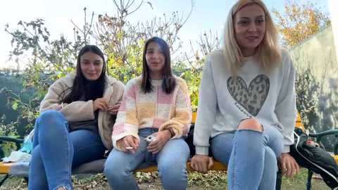 Media: Video of three women sitting on a wooden bench in a garden, wearing casual clothes. One wears a striped sweater, another a beige coat, and the third a white sweater with a heart design. Background features greenery and a fence.