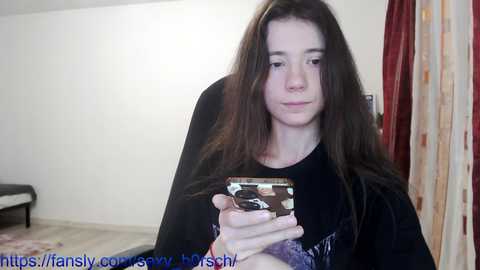 Media: A video of a young woman with long brown hair, wearing a black sweater, holding a smartphone, in a dimly lit room with beige walls and a red curtain.