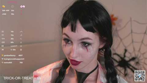 Media: Video of a pale-skinned woman with dark hair in pigtails, wearing a black choker and red lipstick, looking sinister with black makeup around her eyes, in a Halloween-themed room with spider web decorations.