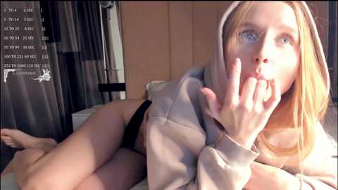 Media: A video of a young, fair-skinned woman with long blonde hair, wearing a grey hoodie and black underwear, lying on a bed, looking up with a finger to her lips.