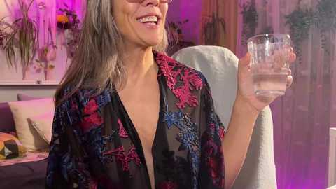 Media: Video of a smiling woman with grey hair, wearing a black floral robe, holding a glass of water. Background features a couch with cushions, floral arrangements, and a purple-lit room.