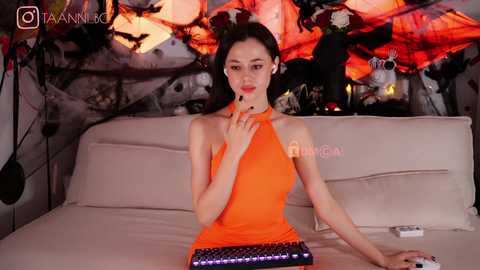 Media: A video of a slender, light-skinned Asian woman with long black hair, wearing an orange halter dress, seated on a beige couch, in a dimly lit room with abstract art and a TV playing.