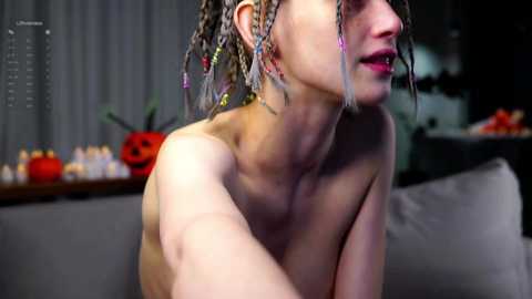 Media: Video of a topless, fair-skinned woman with braided hair, wearing earrings, leaning forward, set in a dimly lit room with a gray couch, a table with candles and a red pumpkin, and a gray curtain.