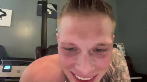 Media: Video of a smiling, tattooed man with short blonde hair, wearing a sleeveless shirt, in an office setting with grey walls and black-framed art.