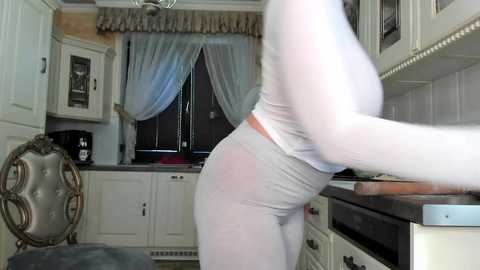 Media: Video of a curvy woman in a sheer white bodysuit, leaning over a kitchen counter, revealing her ample breasts and buttocks. Background features white cabinets, a window with blue drapes, and a silver chandelier.