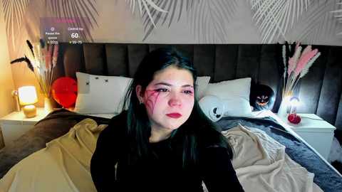 Media: Video of a young woman with dark hair, red lipstick, sitting on a bed with white pillows, surrounded by a cozy, modern room with a gray headboard, white nightstand, and potted plants.