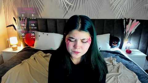 Media: Video of a young woman with long, dark hair, wearing a black shirt, seated on a bed with a gray headboard. She has red makeup on her face. The room has a modern, minimalist decor with a white wall, a nightstand, and a lamp.