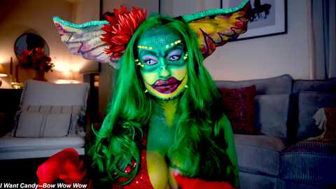 Media: Video of a person dressed as a vibrant, green-skinned, feline creature with large, colorful ears, wearing a red dress, standing in a dimly lit living room with a gray sofa.