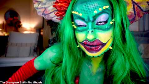 Media: Video of a vibrant green-skinned woman with dramatic makeup, wearing a feathered headdress, red arm bands, and a colorful costume.