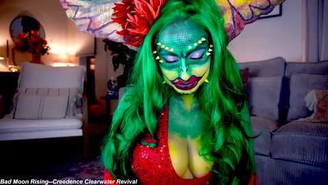 Media: Video of a person in vibrant green makeup with large butterfly wings, wearing a red sequined dress. The background features a cozy living room with a grey couch, white pillows, and a lit lamp.