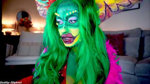 Media: Video of a person with vibrant green makeup, wearing a colorful feathered headdress, sitting on a couch in a dimly lit living room.