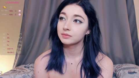 Media: Video of a young woman with pale skin and long, straight, dark blue hair, wearing black earrings. She gazes thoughtfully, standing in a bedroom with gray curtains and a patterned bedspread.