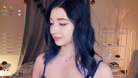 Media: Video of a young woman with fair skin and dark blue hair, wearing a black tank top, standing in a cozy room with a mirror, a white shelf, and a gray curtain.