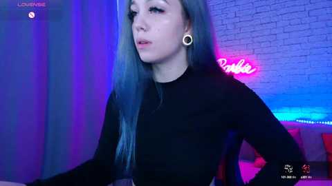 Media: Video of a young woman with long, blue hair, wearing black clothing, and ear gauges, sitting in a dimly lit room with neon lights and a brick wall in the background.