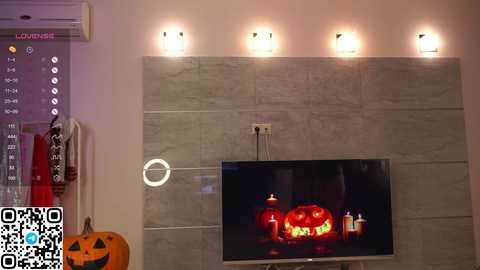 Media: A video of a modern living room with a large, flat-screen TV displaying a jack-o'-lantern image. The wall features a sleek, tiled design with recessed lights. A decorative pumpkin is visible on the left.