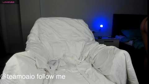 Media: Video of a messy, unmade bed with white sheets, in a dimly lit room with a blue-lit TV in the background. Text at the bottom reads, \"@teammoelio follow me.\