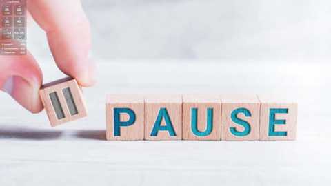 Media: Video of a hand holding a wooden block with the word \"PAUSE\" in blue letters. The block is being placed on a light-colored, textured surface. The background is blurred, emphasizing the wooden block.