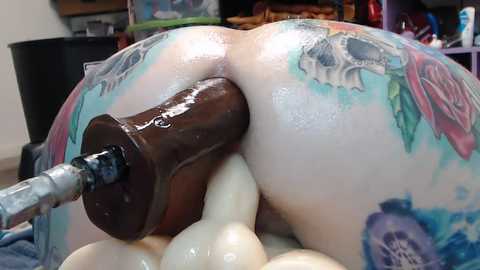 Media: Video of a person's buttocks with a large, shiny, brown dildo inserted anally, covered in colorful tattoos, in a dimly lit room with shelves in the background.