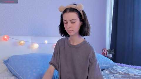 Media: Video of a young woman with short black hair wearing bear ears, gray shirt, and blue bedding, surrounded by fairy lights, in a softly lit bedroom.