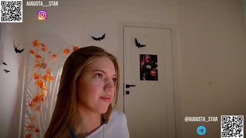 Media: Video of a young woman with long blonde hair, wearing a white top, standing in a room with Halloween decorations and a door with a poster.