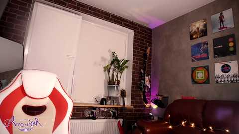 Media: Video of a cozy, modern living room with red brick wall, white window blinds, fern plant, vibrant artwork, red gaming chair, and string lights.