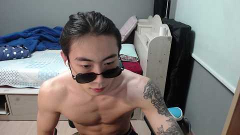 Media: Video of a shirtless Asian man with long black hair, wearing sunglasses and a tattoo on his arm, kneeling in a cluttered, dimly-lit bedroom with a bed and scattered clothes.