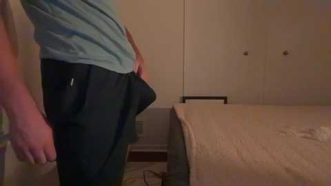 Media: A video shows a person in a blue shirt and black pants, standing beside a bed with beige bedding and white walls in a dimly lit room.