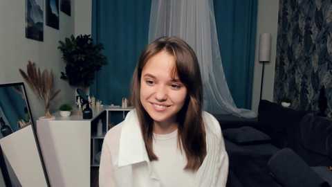 Media: Video of a young, light-skinned girl with straight brown hair, smiling, wearing a white blouse, in a cozy living room with teal curtains, plants, and a dark sofa.