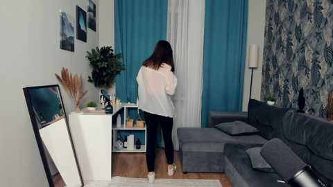 Media: Video of a woman with long brown hair, wearing a white blouse and black pants, standing near a gray sectional sofa in a modern living room with turquoise curtains and blue floral wallpaper.