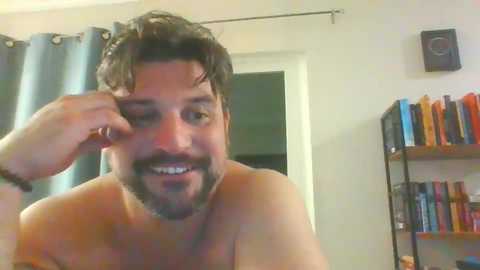 Media: Video of a shirtless man with dark hair and beard, smiling, adjusting his hair, in a room with bookshelves and curtains.