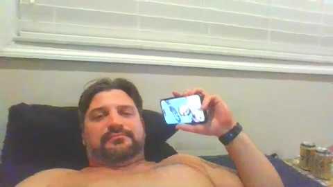 Media: Video of a shirtless, middle-aged man with a beard, lying on a bed, holding a smartphone showing a video. Background includes a white window blind and a nightstand with a glass and a bottle.