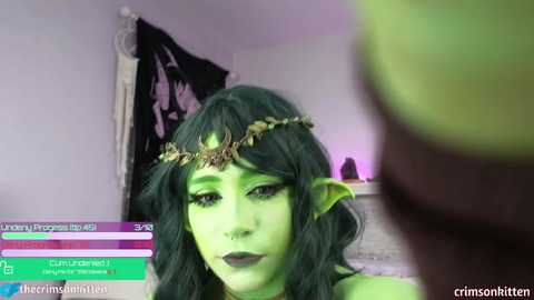 Media: A video of a woman with green skin, wearing a leaf crown and dark makeup, in a dimly lit room. Overlayed is a social media notification.