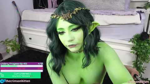 Media: Video of a green-skinned woman with dark green hair and elf ears, wearing a leaf crown, posing topless in a bedroom with a white bed, potted plants, and a gaming interface overlay.