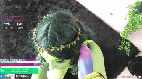 Media: Video of a person with green hair, wearing a green and purple bodysuit, and a leaf crown, lying on a black furry rug with a white wall and green plant in the background.