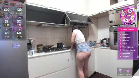 Media: Video of a curvy woman with dark hair, wearing a white crop top and denim shorts, cooking in a modern kitchen with white cabinets and a black stove.