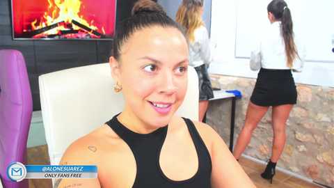 Media: Video of a smiling Latina woman with dark hair in a bun, wearing a black tank top, standing in a modern living room with a fireplace, exposed brick wall, and two other women in the background.