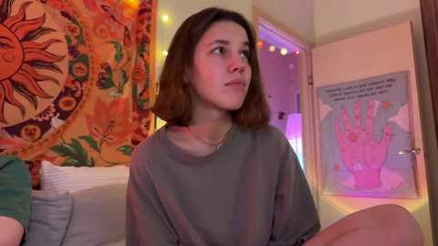 Media: Video of a young woman with shoulder-length brown hair, wearing a grey shirt, sitting on a bed with a sunflower tapestry and a pink hand poster in the background.