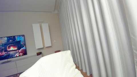 Media: Video of a modern hotel room with white curtains, a flat-screen TV showing a colorful image, and a neatly made bed with white linens.