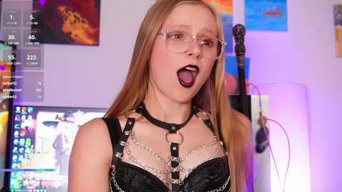 Media: Video of a young woman with long blonde hair, wearing glasses and a black leather harness, open-mouthed, in a vibrant room with colorful posters and a guitar.