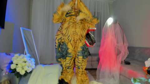 Media: Video of a person dressed in a detailed, colorful, and ornate costume resembling a mythical creature with fur and feathers, standing in a modern living room with white curtains, flowers, and a pink tulle dress in the background.