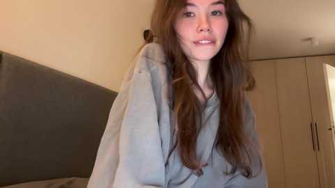 Media: Video of a young woman with long brown hair, wearing a light gray hoodie, smiling softly, in a simple, beige-colored room with a wooden wardrobe in the background.