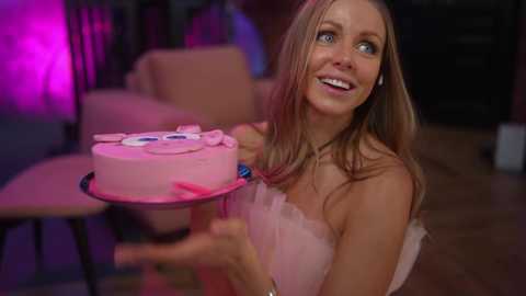 Media: Video of a smiling, light-skinned woman with long, blonde hair, wearing a pink strapless dress, holding a pink cake with a smiling face on it. The background is dimly lit with purple lighting, suggesting a party atmosphere.