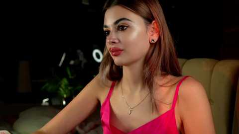 Media: Video of a young woman with fair skin, long brown hair, wearing a bright pink spaghetti strap dress, seated indoors, looking thoughtful.