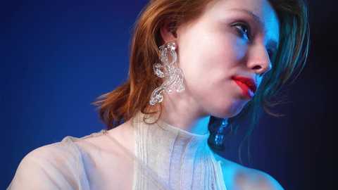 Media: Video of a fair-skinned woman with shoulder-length, auburn hair, wearing a sheer white blouse, and large, intricate crystal earrings, set against a deep blue background, with blue and red lighting.