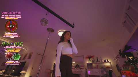 Media: A video of a slender woman with long hair in a white crop top and black high-waisted pants, standing in a dimly lit, modern living room with neon signs and a disco ball.