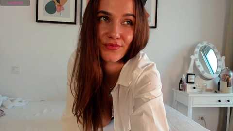 Media: Video of a young woman with long, straight brown hair, wearing a white shirt, smiling softly. Background includes a white vanity table, mirror, and framed art on a white wall.