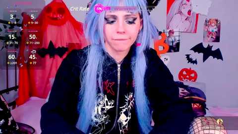 Media: Video of a pale-skinned woman with long, blue hair wearing a black jacket with a graphic design, sitting in a bedroom with Halloween decorations including bats and pumpkins.